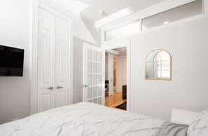 Un dormitorio con cama, televisor, paredes blancas y ventana.