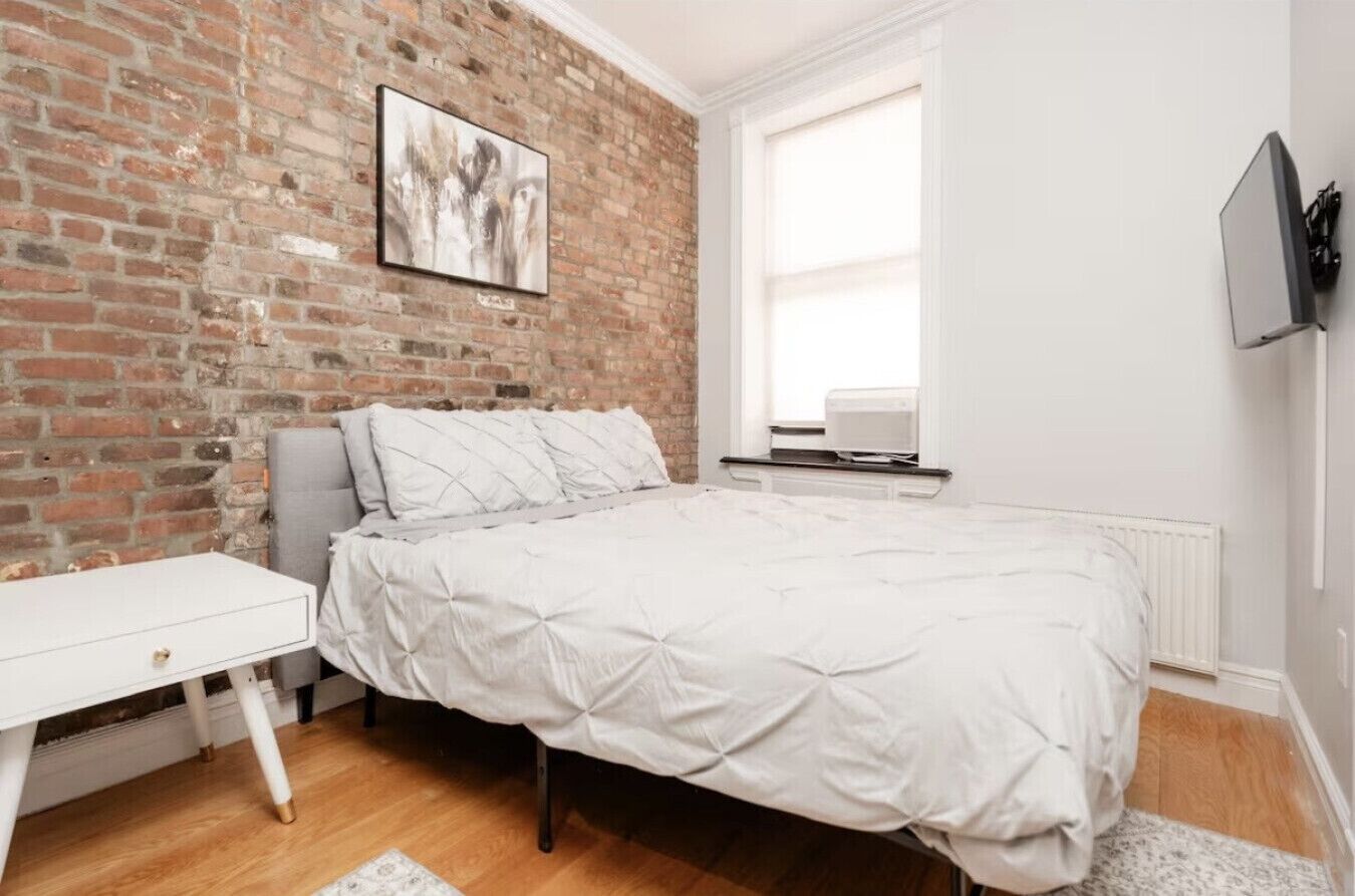Une chambre avec un mur en briques, un lit avec une literie blanche, un bureau blanc et une fenêtre.