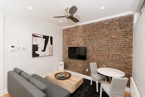 A modern living room with a brick wall, a flat-screen TV, a gray sofa, a coffee table, and a dining area with a chair and table.