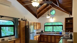 A compact kitchen with wooden cabinets, a dining table with chairs, and a window with a view of greenery. - Villa Tortuga Nosara (Playa Guiones)