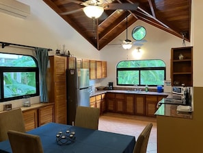 A compact kitchen with wooden cabinets, a dining table with chairs, and a window with a view of greenery.