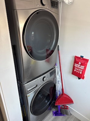 Een gestapelde wasmachine en droger, met een rode branddeken en een bezem op de vloer.