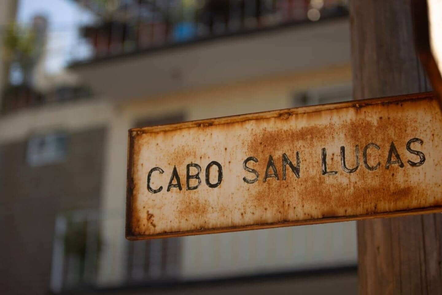 Un panneau de rue usé par les intempéries, portant l’inscription « CABO SAN LUCAS », fixé sur un poteau en bois.