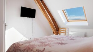 A bedroom with a sloped ceiling, a mounted TV, a window, and a bed with a floral bedspread. - Gîtes de France® - Authenticity and modernity (CUVERVILLE SUR YERES)