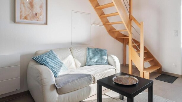 A living room with a sofa, a coffee table, a framed artwork, and a wooden staircase. - Gîtes de France® - Authenticity and modernity (CUVERVILLE SUR YERES)