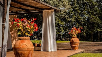 Ett takförsett utomhusområde med terrakottafärgade blomkrukor som innehåller röda blommor, ett vitt draperi och en träpergola.