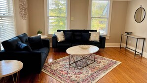 A living room with a black sofa, a round coffee table, a patterned rug, and two windows with blinds.