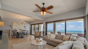 A spacious living area with a ceiling fan, a dining table with chairs, a sofa, and large windows offering a view of the ocean. - Oceans Edge Resort & Spa (San Pedro)