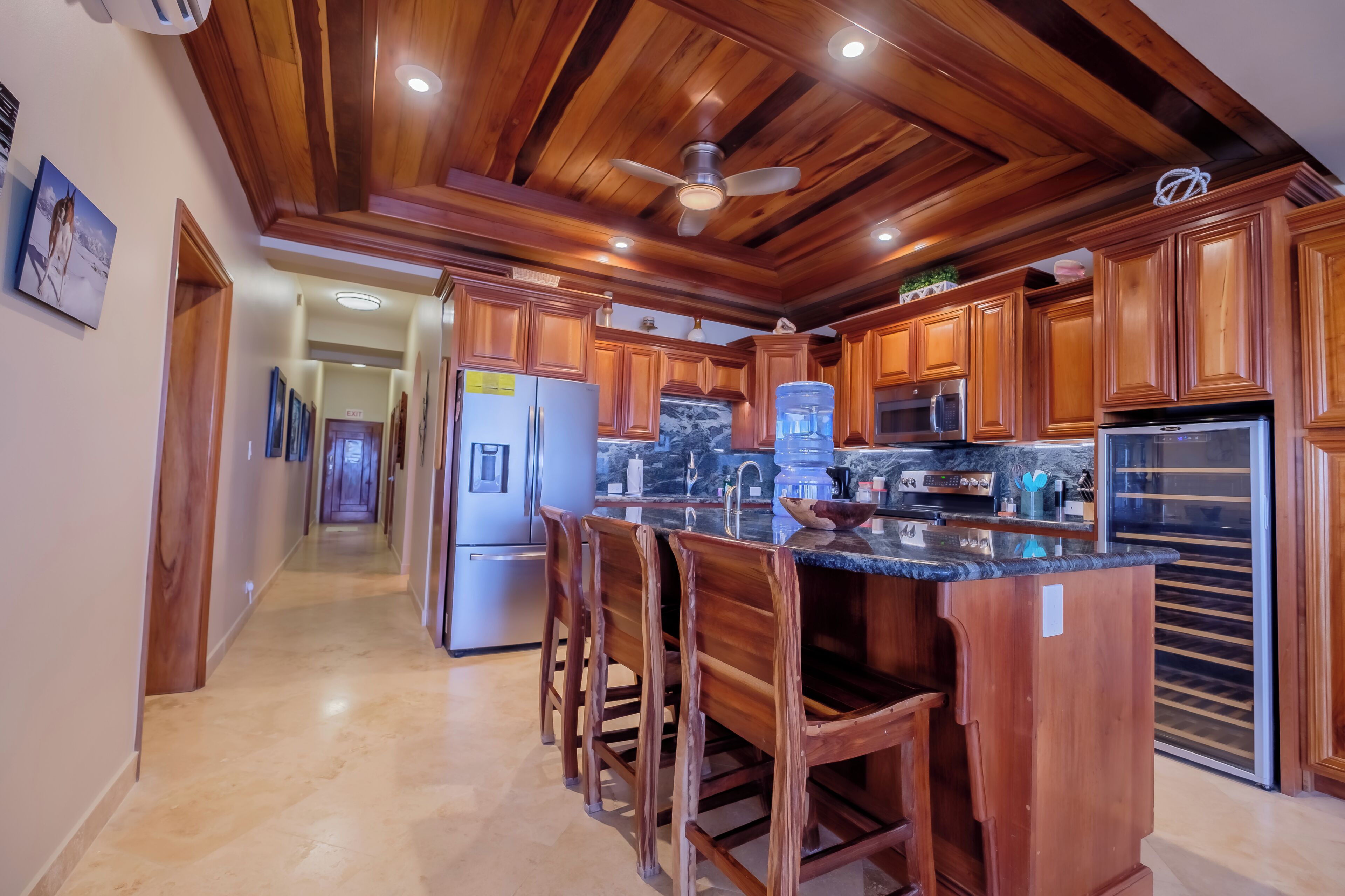A kitchen with wooden cabinets, a central island with bar stools, a refrigerator, a microwave, and a water dispenser.