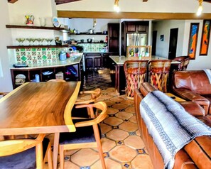 Zona pranzo con tavolo e sedie in legno, una panca con una coperta e una cucina sullo sfondo.