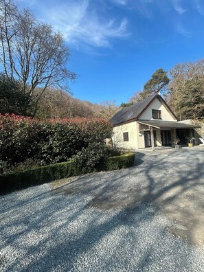 A house with a gravel driveway, a covered porch, and a garden with bushes and trees. - Mawddach View in Snowdonia with Hot tub, pet friendly & great location (Barmouth)