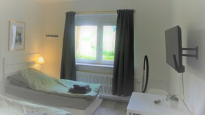 A hotel room with a bed, a desk, a chair, a TV, a window with curtains, a radiator, and a picture on the wall. - Work Inn Rhein Ruhr Apartment (Duisburg)