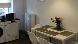 A compact kitchen with a washing machine, a dining table with chairs, a hanging light fixture, and a radiator. - Work Inn Rhein Ruhr Apartment (Duisburg)