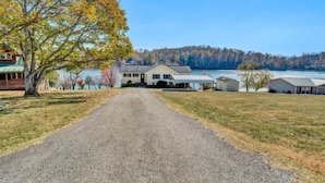A gravel driveway leading to a house with a lake view, surrounded by trees and additional buildings. - Lakefront Escape: hot tub, endless views, deep water dock, gentle walk to water (Goodview)