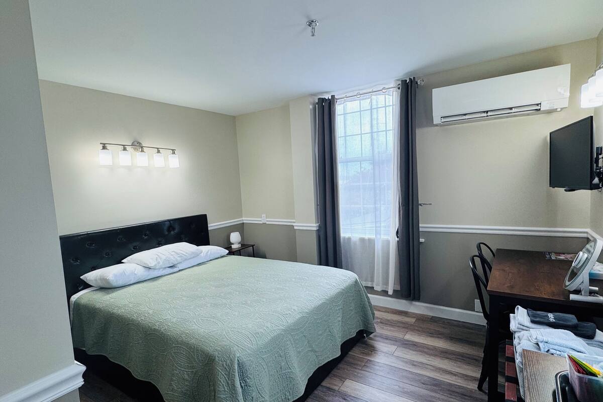 Una camera d'albergo con un letto, una scrivania, una sedia, una finestra con tende, un condizionatore d'aria e una TV fissata al muro.