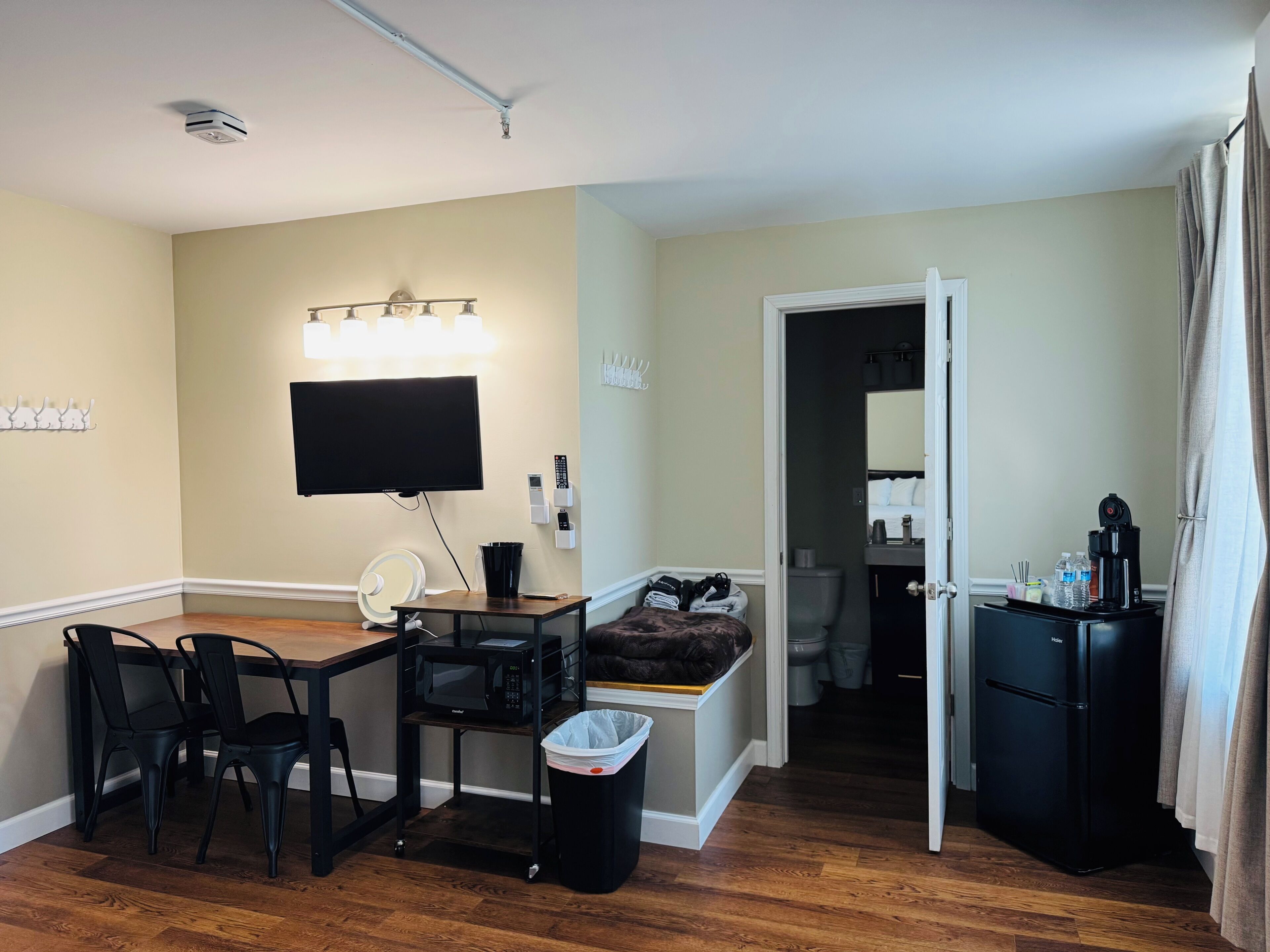 A compact hotel room with a dining area, a television, a microwave, a trash bin, a small table with a chair, and a refrigerator.