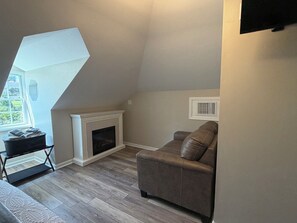 A room with a sloped ceiling, a fireplace, a brown leather sofa, and a window with a view of greenery.