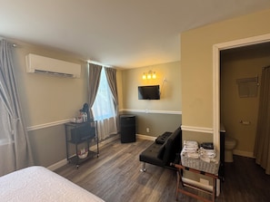 Une chambre dâhĂŽtel avec un lit, un bureau Ă©quipĂ© dâun ordinateur, un tĂ©lĂ©viseur fixĂ© au mur, une poubelle et une salle de bain visible Ă travers une porte ouverte.