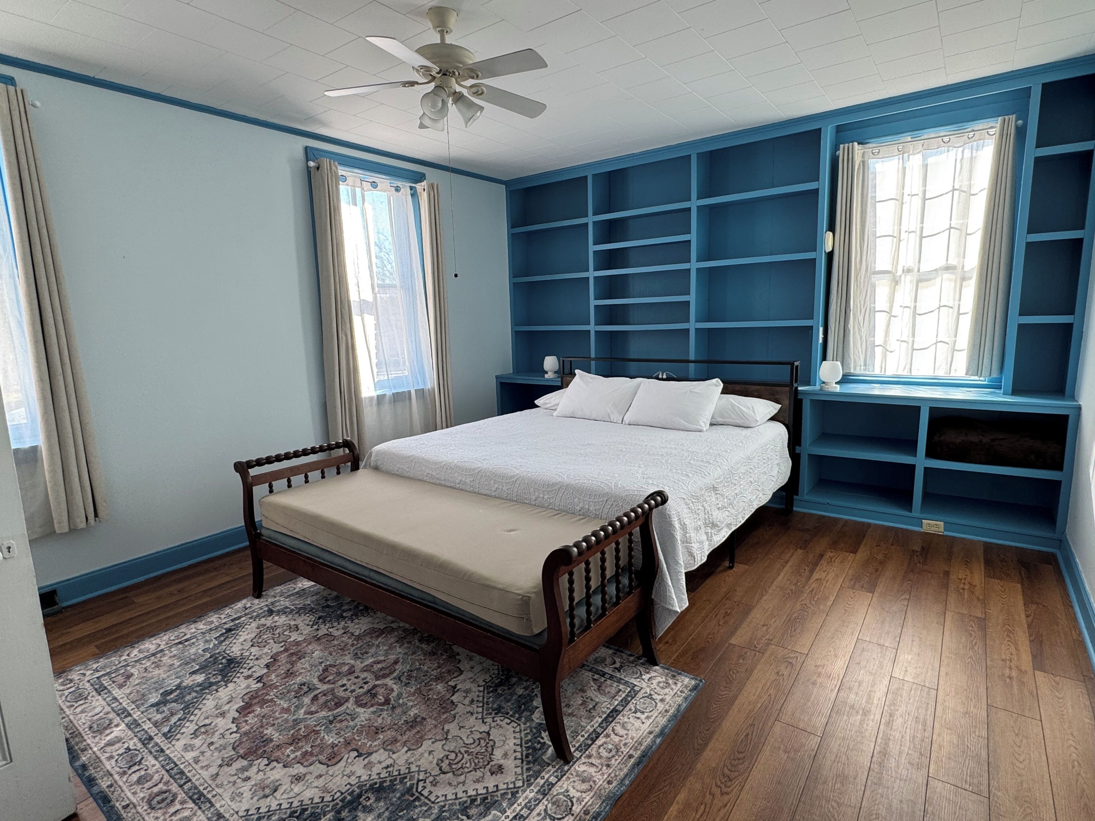 Un dormitorio con cama, mesita de noche, ventana con cortinas, ventilador de techo y estanterías empotradas.