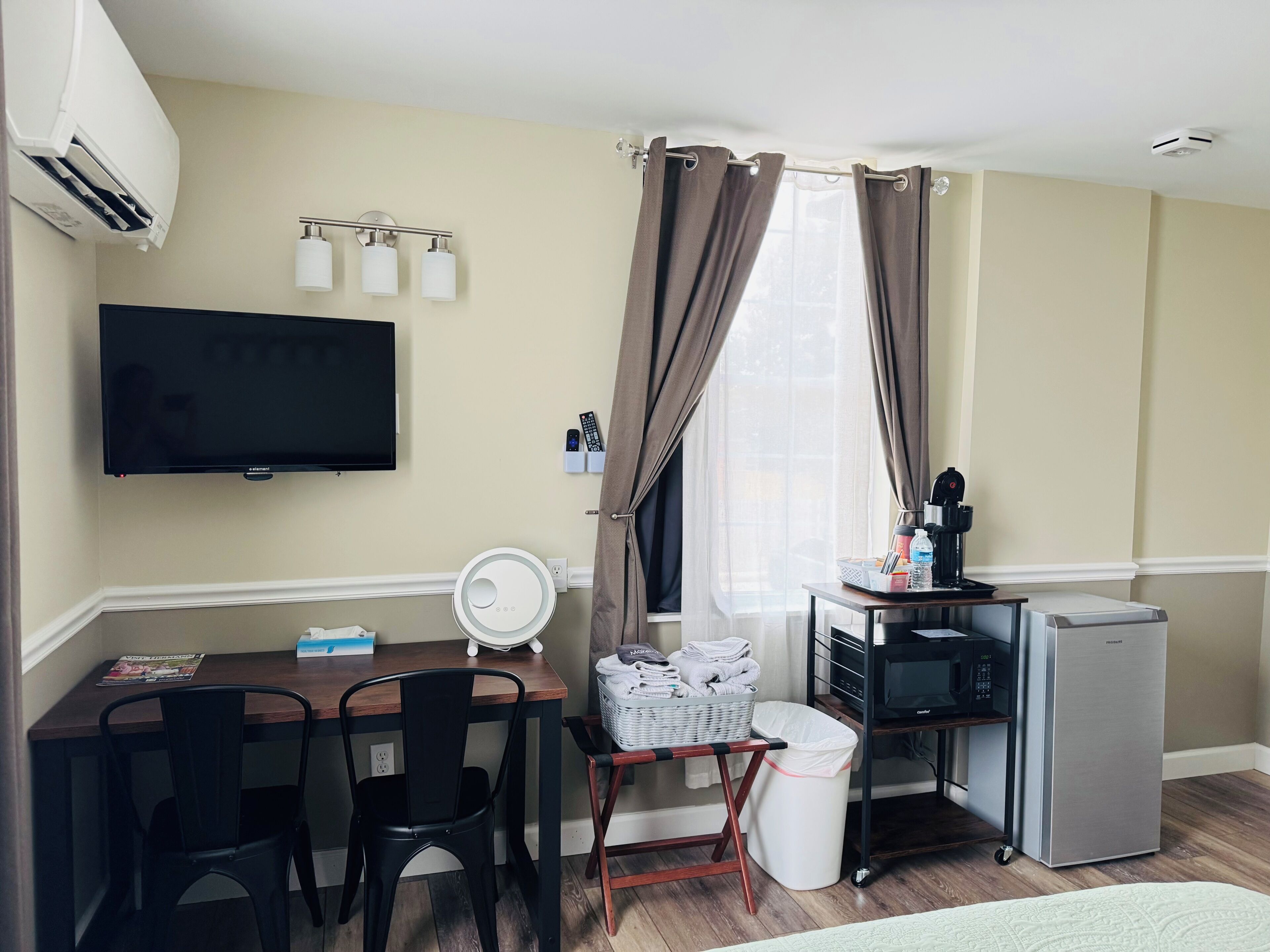 A compact hotel room with a flat-screen TV, a desk with a chair, a small kitchenette, and a window with curtains.