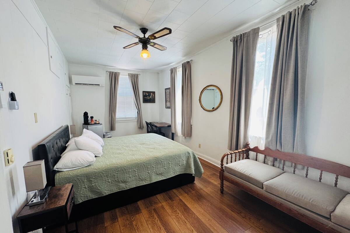 Una camera da letto con un letto, pavimento in legno, ventilatore a soffitto, comodino, tavolo da notte, una finestra con tende, uno specchio e un divano.