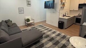 Ein modernes Wohnzimmer mit einem grauen Sofa, einem kleinen weißen Sessel, einem gemusterten Teppich und einem an der Wand befestigten Fernseher.