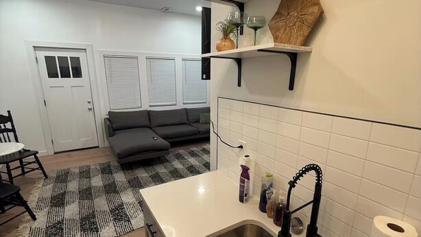 Eine moderne Küche mit Spüle aus Edelstahl, weißen Fliesen und einem schwarzen Wasserhahn. Es gibt einen Essbereich mit einem schwarz-weißen Teppich und einem grauen Sofa.