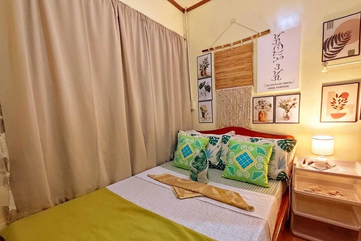 Una cama bien tendida con almohadas estampadas, una lámpara de noche y una pared con cuadros.