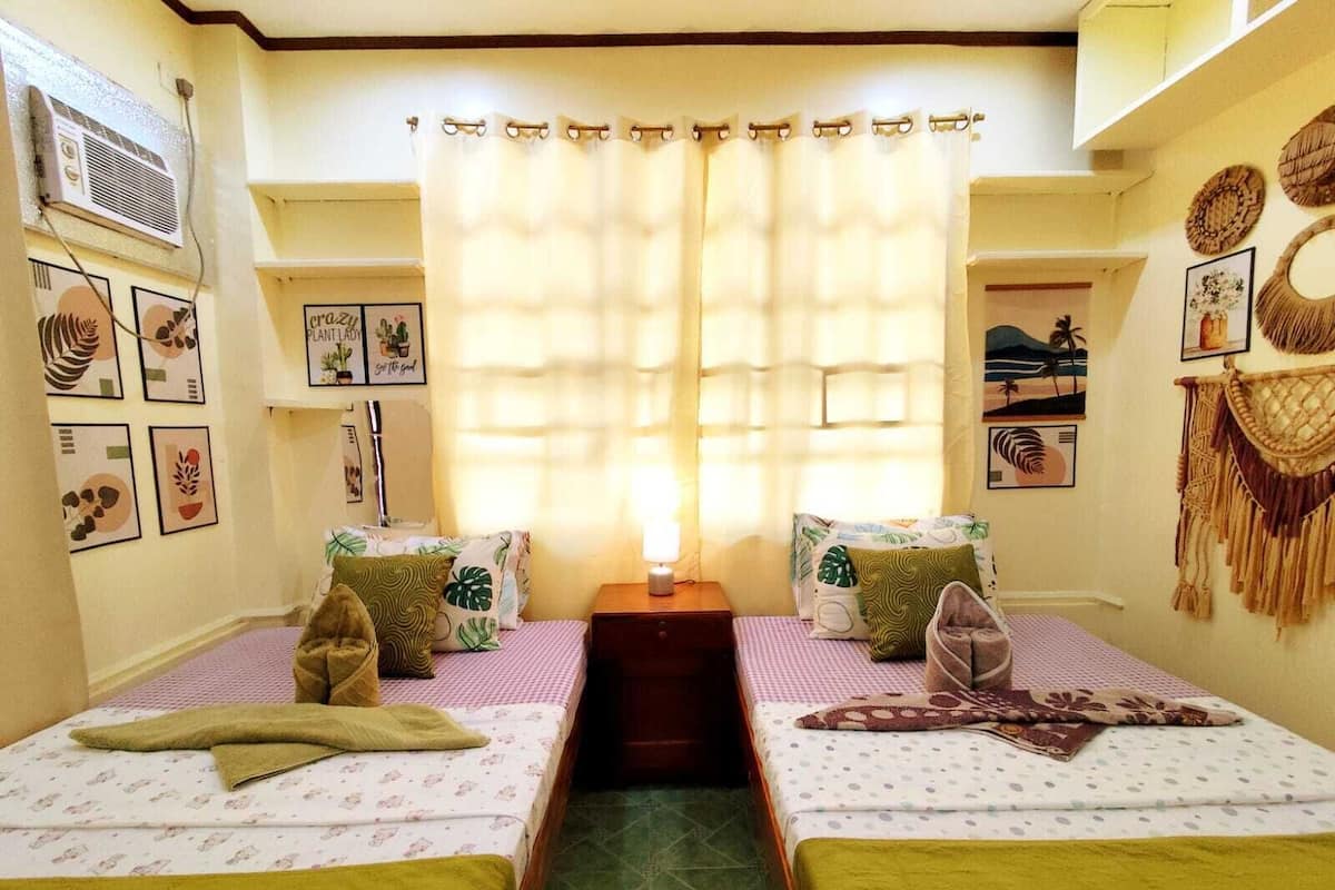 Una habitación con dos camas, una ventana con cortinas, decoración en la pared y una pequeña mesa con lámpara.