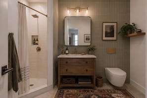 A bathroom with a shower, a sink with a mirror, a toilet, and a shelf with a plant.