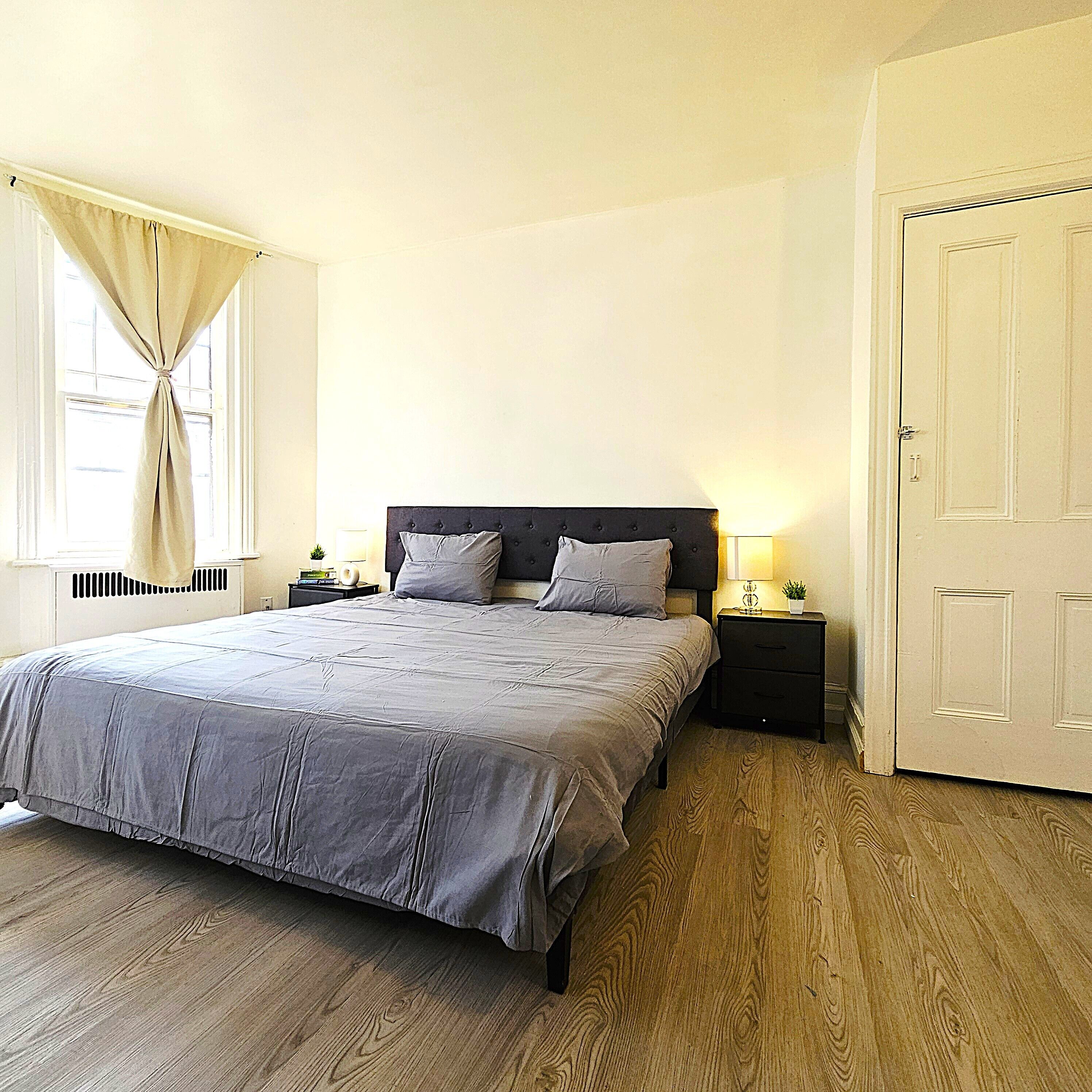 A bedroom with a bed, two bedside tables, a nightstand, a door, and a window with curtains.