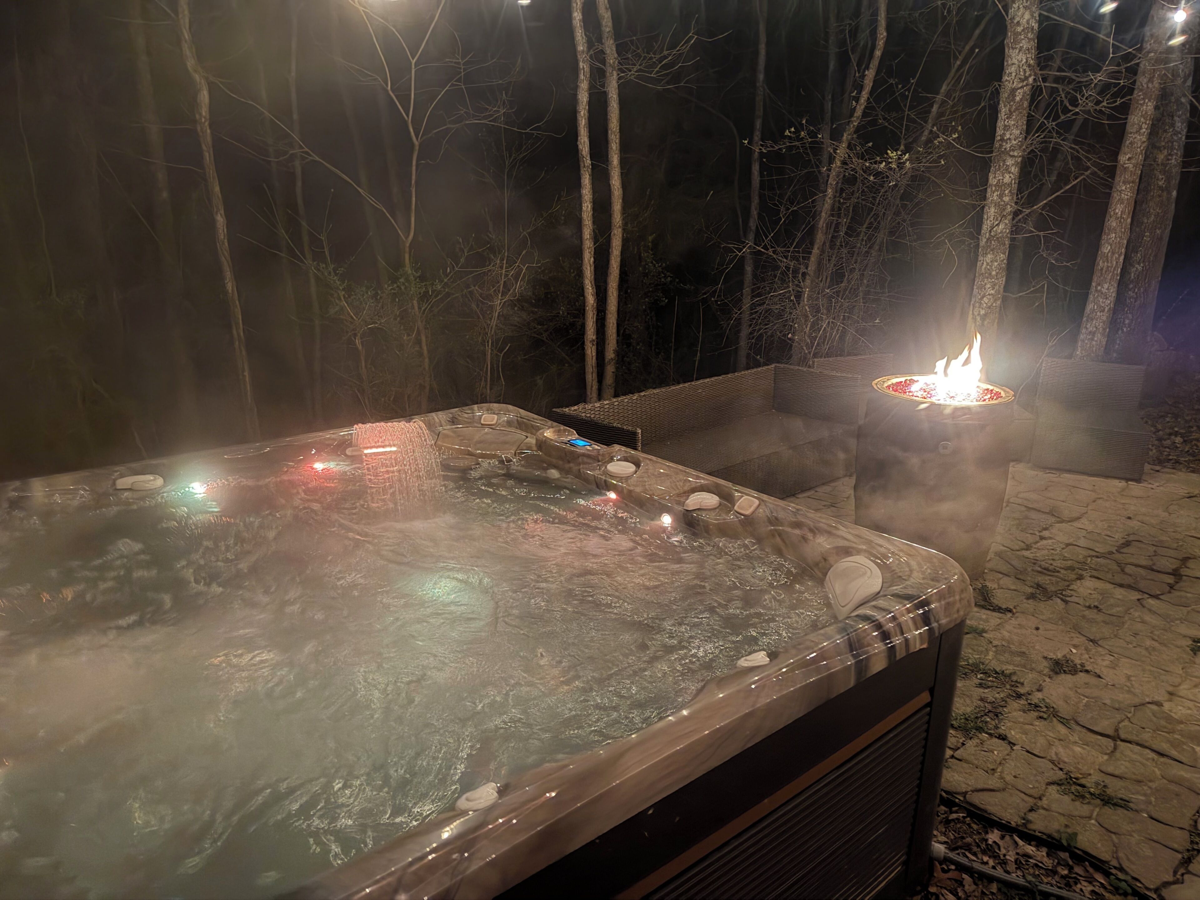 A hot tub with a fire feature, surrounded by trees and a wicker chair.