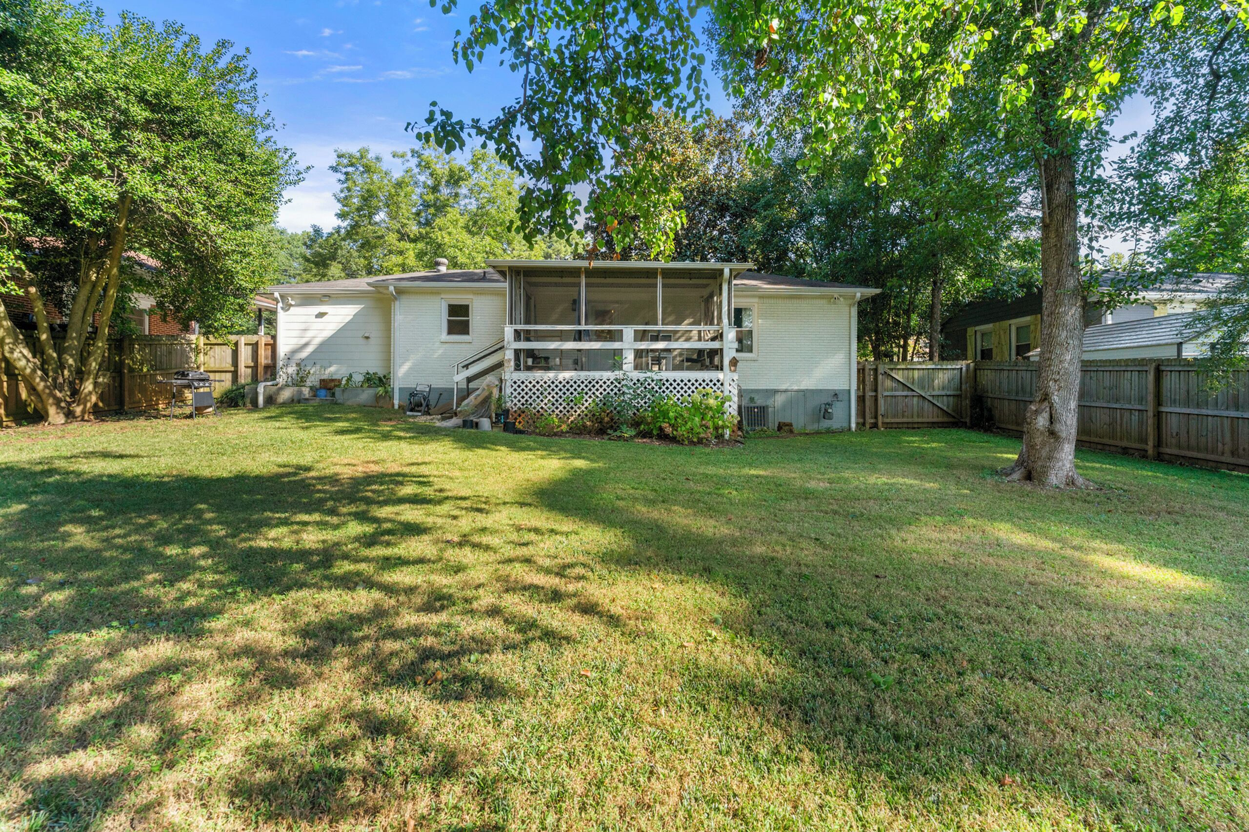 A single-story house with a front porch, a fenced yard, and a large tree in the background.