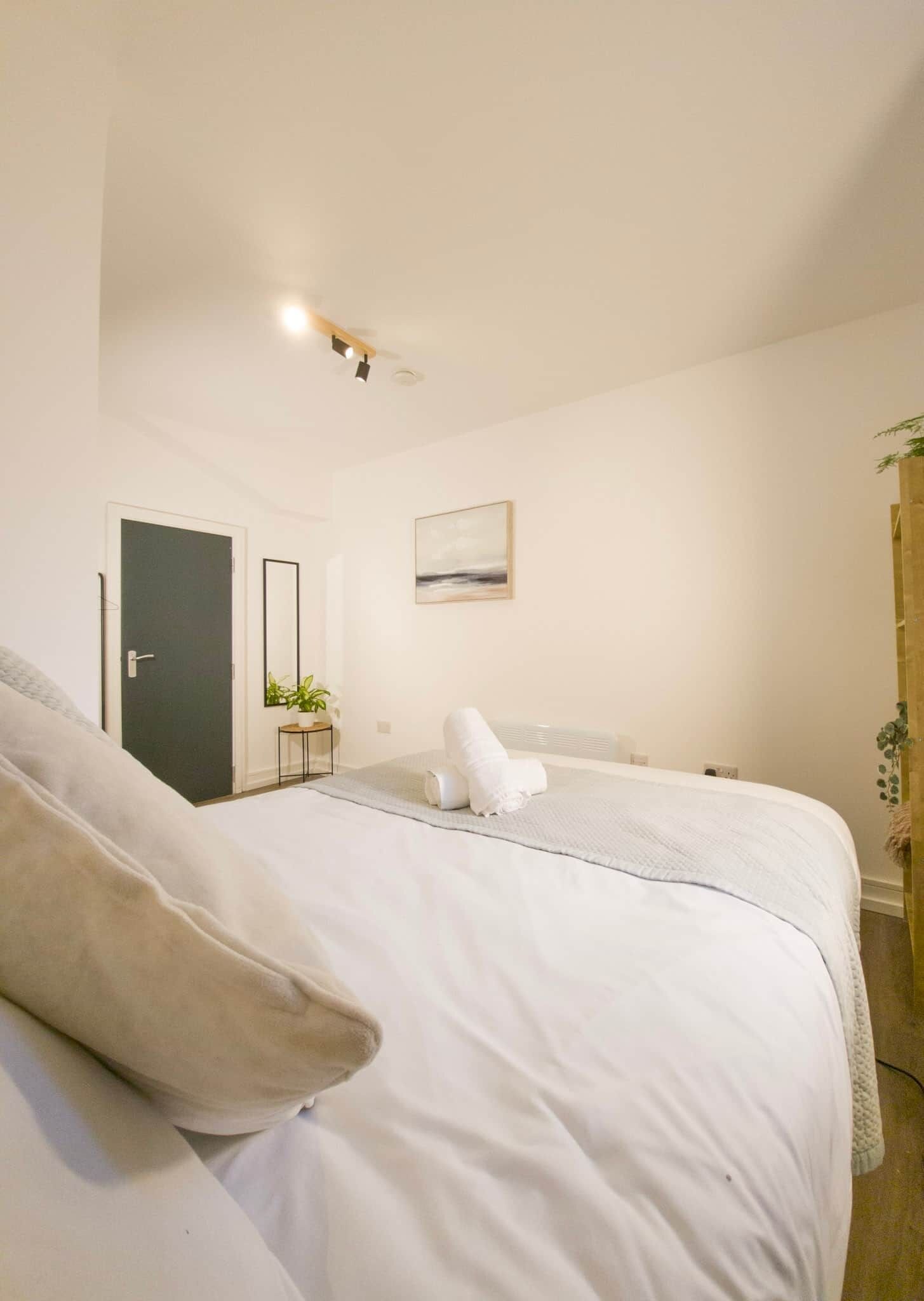 Un dormitorio con cama, una puerta, una mesita con una planta, un cuadro en la pared y una planta en la mesita de noche.