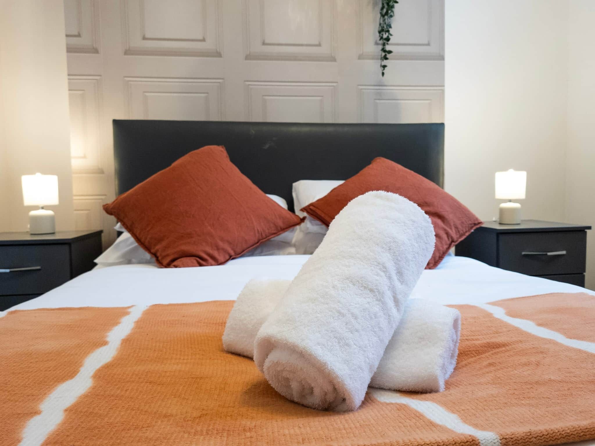 Un lit bien fait, avec des serviettes blanches et orange, deux oreillers rouges et une tête de lit noire.