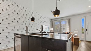 Et moderne køkken med en central ø, hængende lamper og et vindue med persienner.