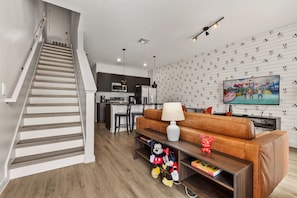 A modern living room with a staircase, a kitchen in the background, a television, and a wall with a Mickey Mouse pattern. - Family 3BR townhouse w/ pool & balcony (Kissimmee)