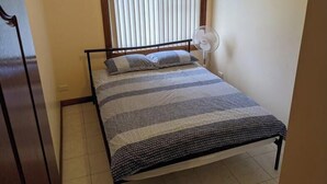 Ein kleines Zimmer mit einem Einzelbett, einem Fenster, einem Standventilator und einer schlichten Wand.