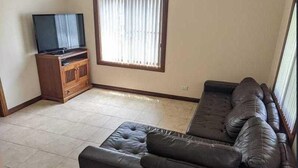 Ein Wohnzimmer mit einer Eckcouch, einem Fernseher auf einem Holzboden und einem großen Fenster mit Jalousien.