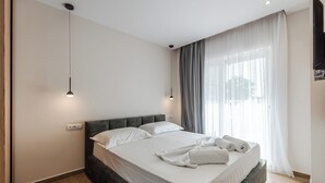 Ein modernes Hotelzimmer mit einem großen Bett, zwei Nachttischlampen, einem Flachbildfernseher und einem Fenster mit durchsichtigen Vorhängen.