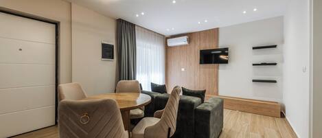 A modern living room with a round dining table, two beige chairs, a dark green sofa, a flat-screen TV mounted on the wall, and recessed ceiling lighting.