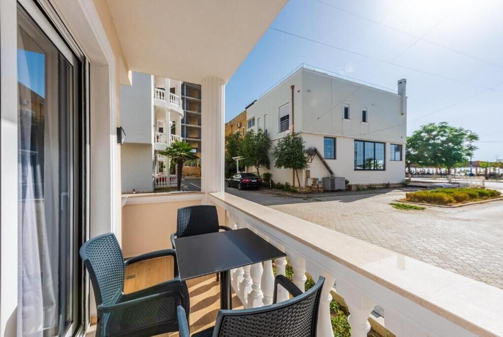 A balcony with a table and chairs, overlooking a street with buildings and a parked car.