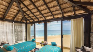 Quarto com vista para a praia, duas camas com roupa de cama estampada, uma mesinha com uma planta e um telhado de palha.