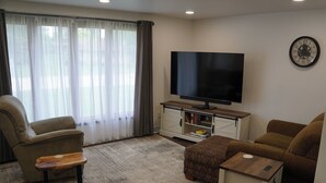 Ein Wohnzimmer mit Flachbildfernseher, einer Couch, einem Sessel, einem Couchtisch und einem Bücherregal.