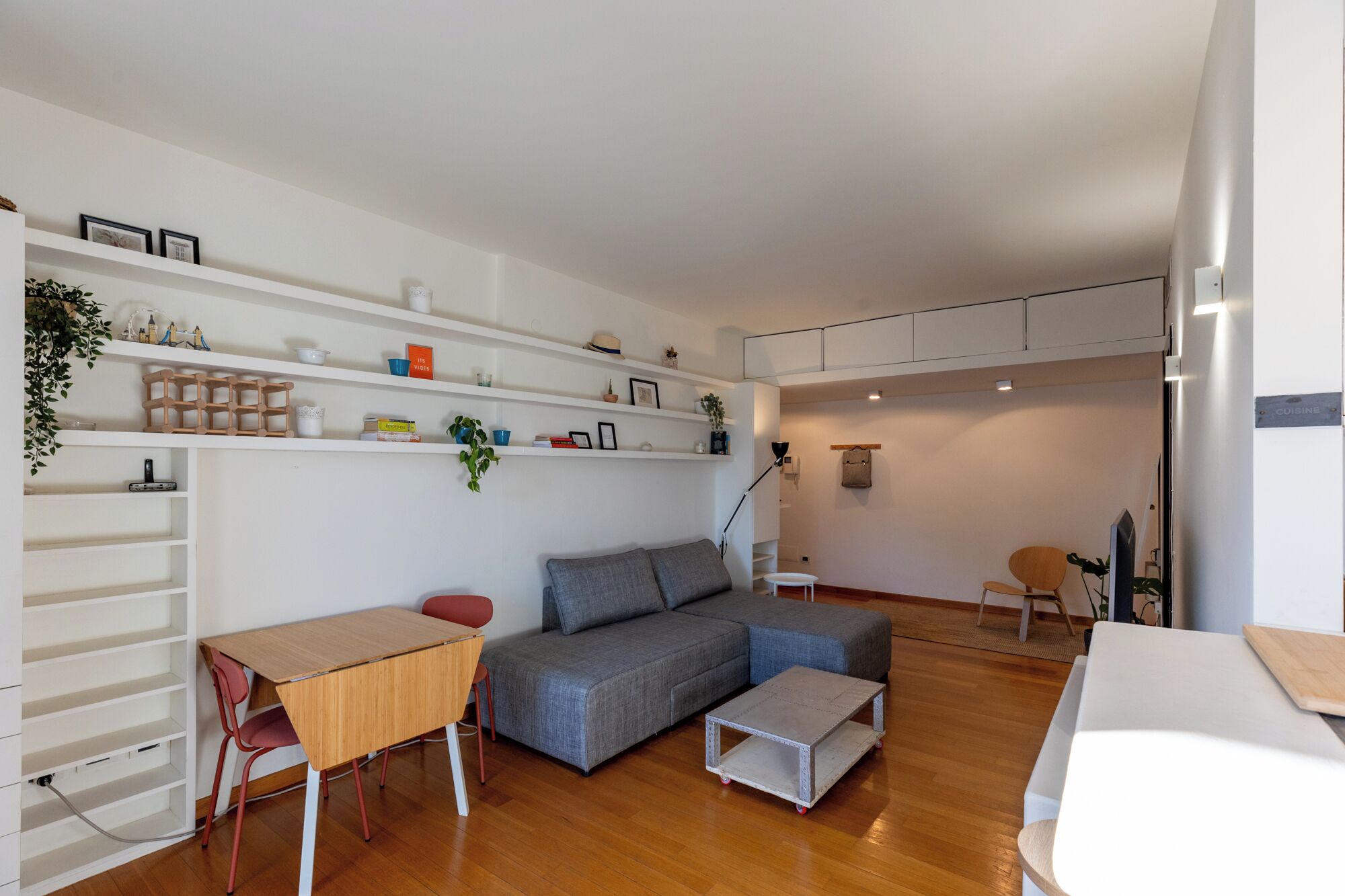 A modern living room with a sofa, a small table, and a TV. There are shelves with various items and a plant.