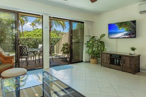 A spacious living area with a glass sliding door leading to a balcony, a flat-screen TV mounted on the wall, and a wooden cabinet. - Maui Kam D106 | Best Location | Spacious Lanai + Lush Lawn Access (Kihei)