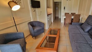 Sala de estar com sofá, duas poltronas, mesa de centro, televisão e um espaço de refeições ao fundo.