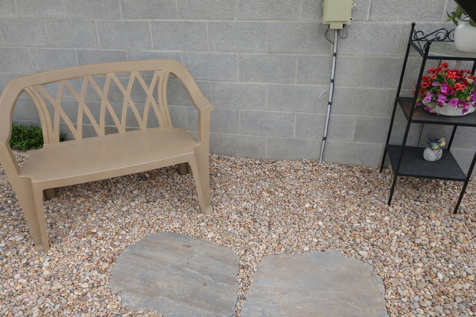 A beige bench, a stone pathway, a wall-mounted electrical box, and a metal shelf with flowers.