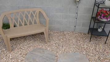 A beige bench, a stone pathway, a wall-mounted electrical box, and a metal shelf with flowers.