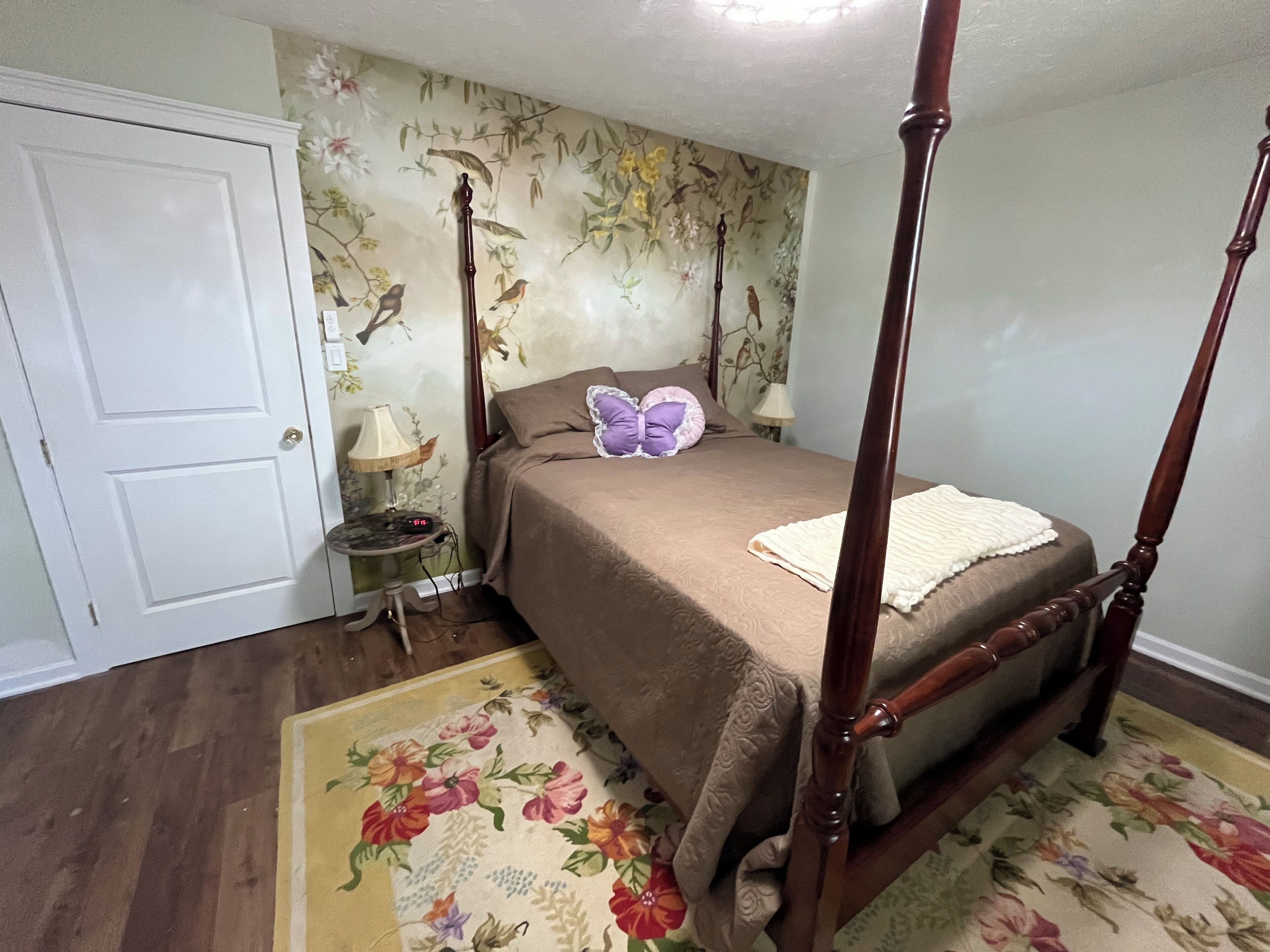 A four-poster bed with a floral-patterned bedspread, a nightstand with a lamp, and a floral rug.